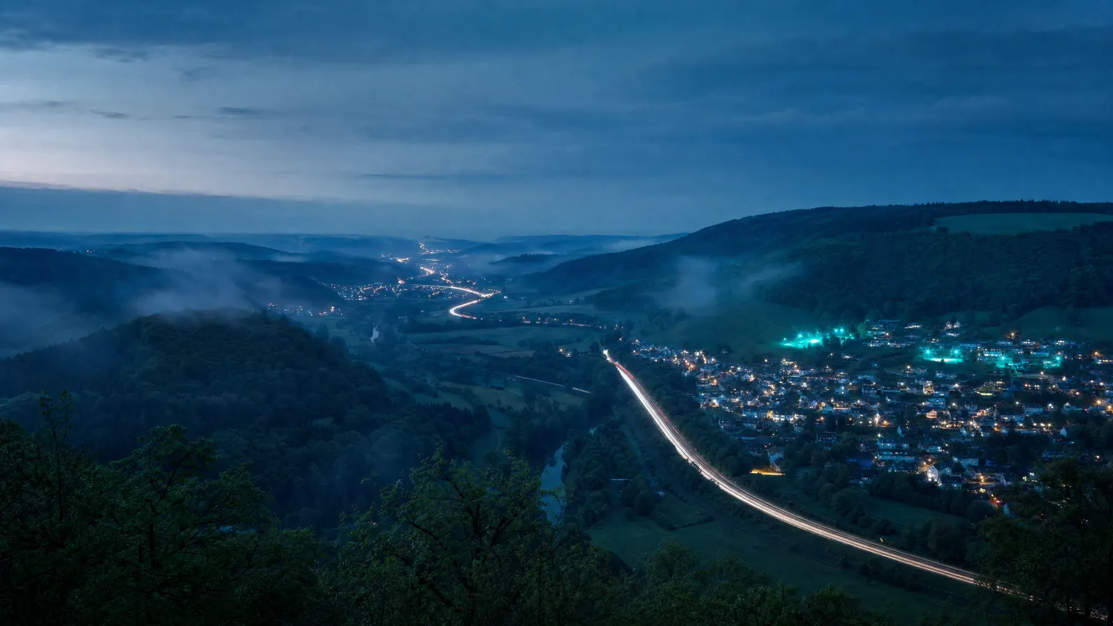 Weites Flusstal mit Dörfern, Autobahn und bewaldeten Hügeln zur blauen Stunde